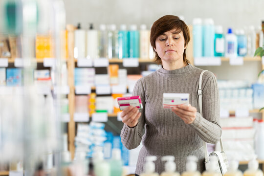 Thoughtful woman carefully comparing two different packs of paracetamol tablets while choosing effective analgesic and antipyretic agent, checking dosage before purchase