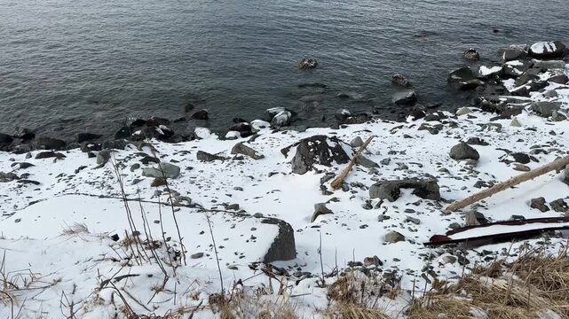 Close up of snow covered rocky ocean shore, high angle looking down fresh white snow at the edge of cold rippling bay waters.
