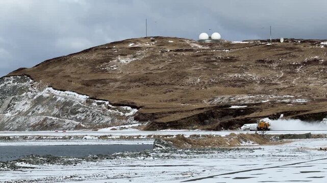 Establishing shot of Tom Madsen Airport showing a snow plow on the runway, with radar domes sitting atop a brown tundra hill, Dutch Harbor.