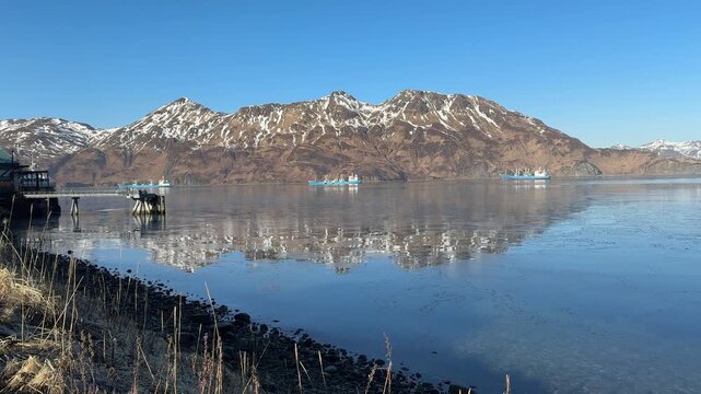 Wide scenic landscape shot across ocean water filled with slush ice sheets and anchored commercial freighter vessels.