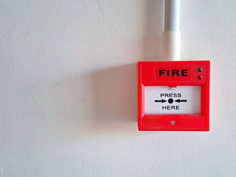 Close-up of a red fire alarm call point with 'PRESS HERE' text mounted on a clean white wall. Safety, emergency equipment, and security stock photo.