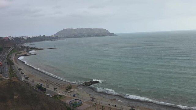 Scenic wide angle view of a coastal highway and ocean beach line under an overcast sky with a hill in the background
