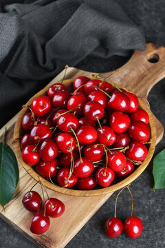Fresh ripe sweet cherries in wooden bowl on rustic cutting board with green leaves and linen cloth on black textured background. Healthy summer fruit, red berries, fresh seasonal harvest, healthy food