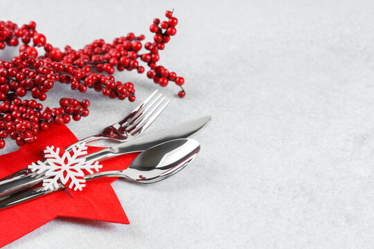Christmas table setting with cutlery arranged in red star shaped cutlery holder decorated with white snowflake, holly berry branch on light gray textured background with copy space