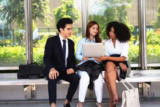 Asian businesswoman holding a laptop while two colleagues look on beside leafy hedges on a bright city business plaza, glass canopy and dense green hedges, cream tote and black briefcase