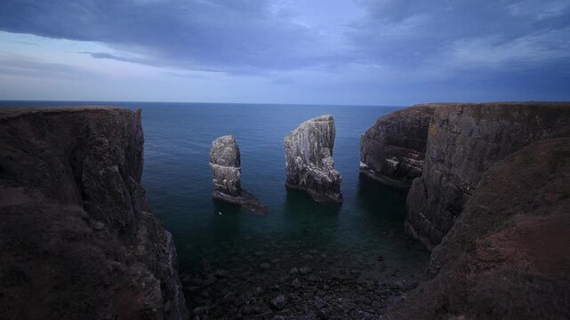 Two towering limestone sea stacks stand between dark coastal cliffs beneath a cloudy evening sky.