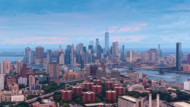 Lower Manhattan skyline across the river from Brooklyn. Towers in New York rise behind bridge spans and brick housing beneath layered clouds and a pale pink horizon.