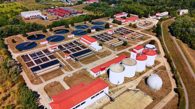 Aerial view of a wastewater treatment plant and storage tanks. Circular basins and rectangular channels surround buildings with red roofs beside trees and service roads.