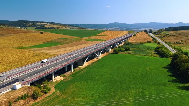 Road bridge extending across fields toward distant hills. A divided carriageway rests on repeated concrete arches above crops and trees in direct sunlight.