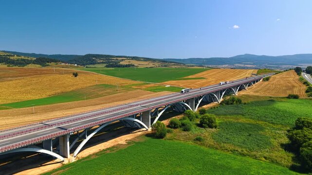 Motorway viaduct running diagonally through farmland. Arched supports cast shadows on green fields as vehicles cross beneath an open blue sky.