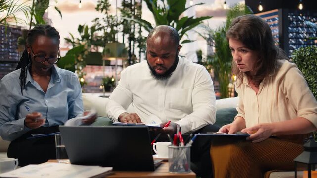 Shocked businesspeople react to low company performance metrics on a laptop during a rooftop meeting, showing negative emotion while reading document charts.