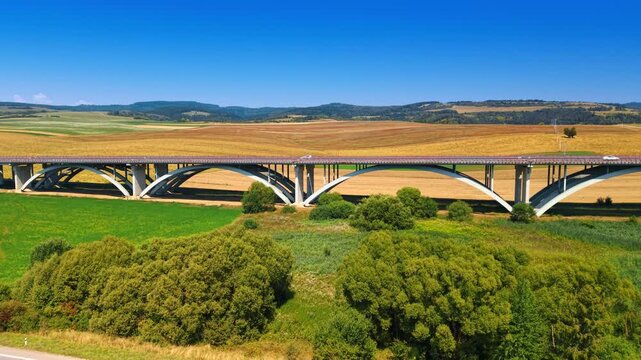 Concrete bridge arches above green fields and shrubs. A side view shows the roadway crossing farmland, with rolling hills beneath a clear blue sky.