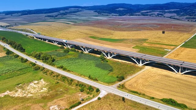 Motorway viaduct and road junction among agricultural fields. An overhead oblique view shows crossing routes, vehicles and concrete arches beside green and brown parcels.