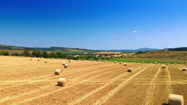 Straw bales and parallel tracks across a harvested field. An elevated view shows cylindrical bundles on golden stalks, with a distant road bridge and hills.