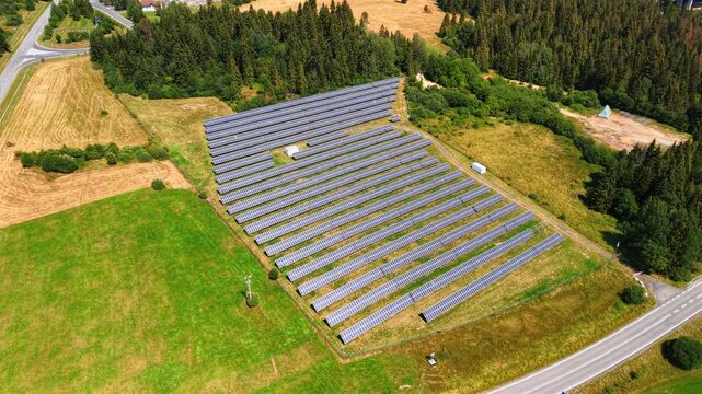 Overhead view of parallel solar panel rows beside farmland. Dark rectangular modules cover a fenced plot between green fields, forest and a paved road.
