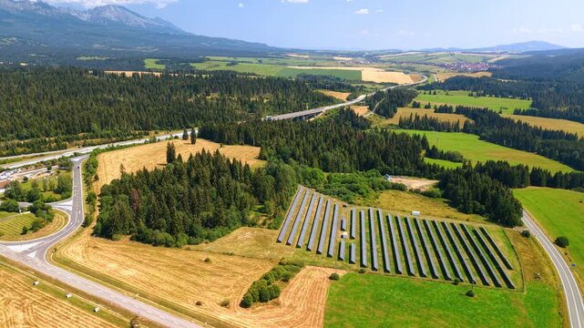 Solar panel array among fields, forest and roads. An aerial view shows parallel dark modules beside golden parcels and green woodland, with hills beyond.
