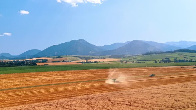 Combine harvester raising dust in a field below mountains. A tractor and trailer follow the crop strips, with green farmland and hazy ridges beyond.