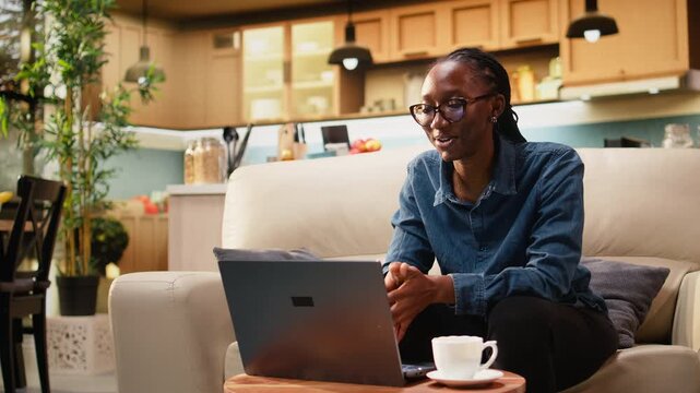 Young black woman using laptop during video call at home, engaged in virtual meeting for remote work. Modern telework lifestyle highlights communication in web conference, multitasking.