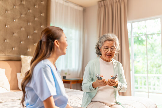 Asian female caregiver or physiotherapist helping senior woman exercise with hand gripper at home. Elderly encouraging strength training, physical activity, healthy aging, mobility and rehabilitation