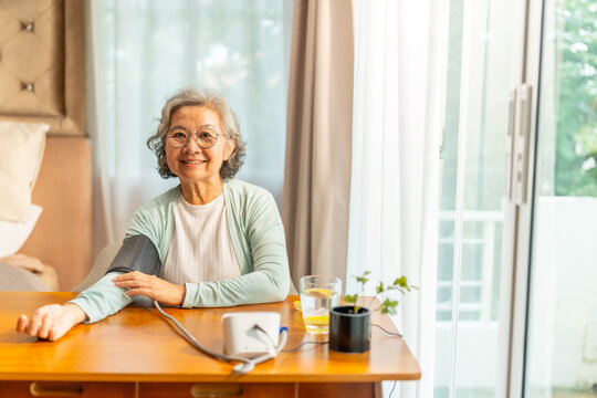Asian senior woman measuring blood pressure at home using digital monitor, elderly person checking hypertension and cardiovascular health during daily healthcare routine and wellness monitoring