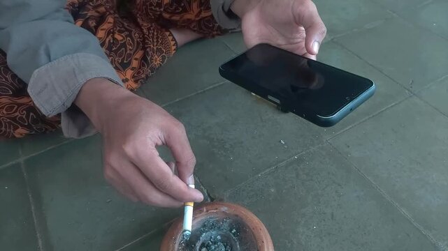 Person's hands smoking cigarette and using smartphone on tiled floor