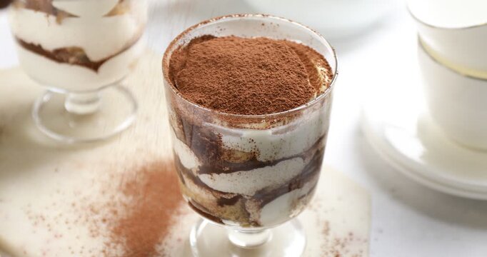 Sprinkling tasty tiramisu in dessert coupe with cocoa powder at white table, closeup
