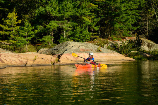 Sea kayaker in an orange kayak paddles past a lichen-covered granite dome and pine forest in morning light on Georgian Bay, Ontario, Canada