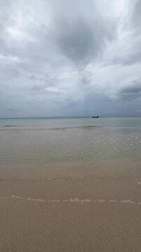 Gentle waves approach the shore under a cloudy sky with distant boats in the ocean