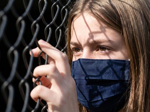 Young person in a protective face mask looking contemplatively through a metal fence, symbolizing feelings of restriction and hope during uncertain times