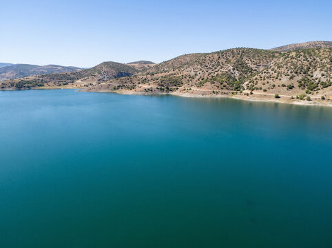 Stunning aerial drone view of a vast turquoise water reservoir surrounded by dry, sun-drenched hills and winding coastal roads under a clear blue sky, capturing a peaceful and serene wild landscape.