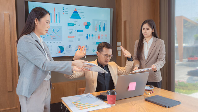 A stressed Asian business team having a tense meeting, with a female manager standing and gesturing while a male colleague looks overwhelmed and holds his head, during a difficult project discussion i