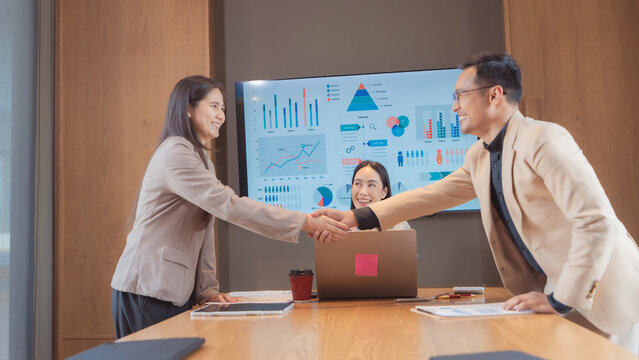 A group of happy Asian business people stacking hands together in the center of a table to show unity, teamwork, and commitment after a successful presentation in a modern office meeting room.