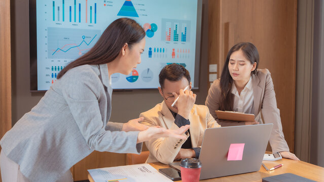 A stressed Asian business team having a tense meeting, with a female manager standing and gesturing while a male colleague looks overwhelmed and holds his head, during a difficult project discussion i