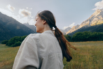 Obraz premium Woman in casual shirt stands in a meadow with mountains in the background, capturing a serene outdoors lifestyle moment in nature, hair tied back and bright sky
