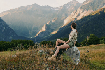 Obraz premium Woman sitting on a rock in a mountain meadow, enjoying outdoor scenery. Natural light highlights the valley and distant peaks, conveying serenity, exploration, and nature connection.