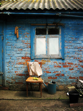 A fragment of a brick village house with peeling blue paint on the walls. An old wooden chair with pears rests under the window. Next to it is a metal watering can. Corn cobs hang on the wall. Rural.