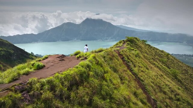 Man hiking on green grass mountain ridge offering an expansive aerial view of Lake Batur and Mount Batur volcano caldera, symbolizing adventure, travel, and exploration on Bali island, Indonesia