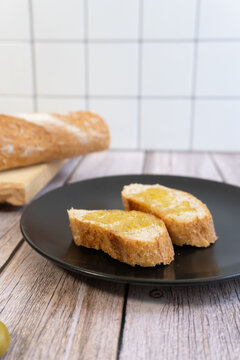 Slices of fresh bread on a black plate. Olive oil is drizzled over each slice. Behind them, on a wooden surface, rests a loaf of bread.