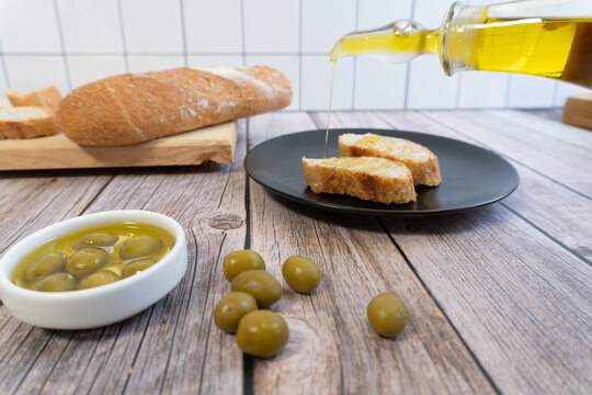 Golden oil drips onto rustic bread slices. Olives rest beside a bowl of green olives. Wooden table holds bread, oil, and olives. Perfect for food, wellness