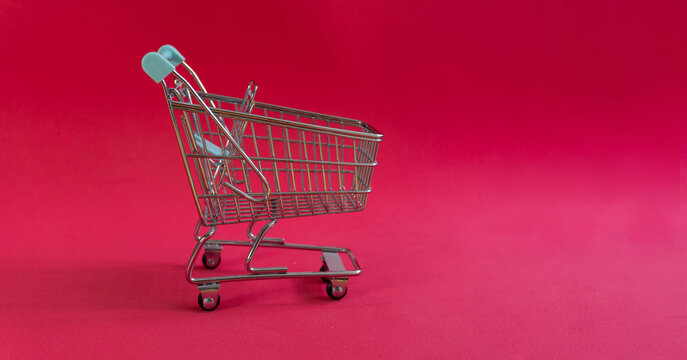 Miniature shopping cart on vibrant red background. Metal frame with blue handle. Modern, clean design perfect for e-commerce themes. Great for digital ads or retail concept visuals