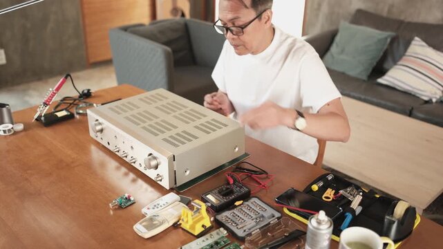 Mature Asian man unscrewing and disassembling audio equipment, checking circuit board for repair, home electrical appliance DIY fix and maintenance, electronics component hobby, handyman workshop.