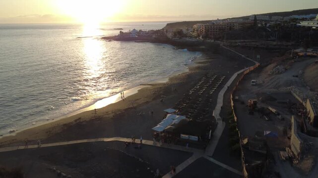 Vista a&eacute;rea de una playa curva de arena negra con tumbonas y mar brillante al atardecer, La Caleta, Costa Adeje, Tenerife