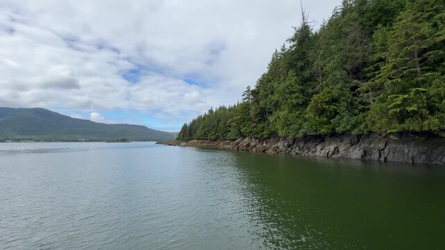 Wide establishing shot from a boat tracking past a calm green bay bordered by a thick temperate rainforest ridge in southeast Alaska. 