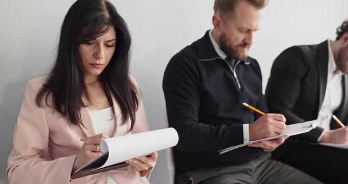 Applicants complete paperwork and application during interview. Seated in a waiting area, a woman and two men write on clipboards, preparing for hiring assessment. Hiring theme is made clear.