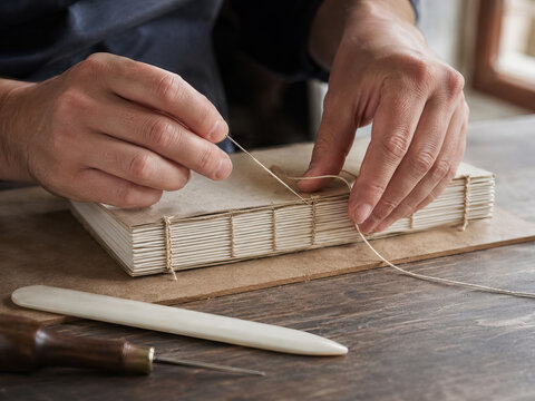 A man meticulously sews together the pages of a handmade book on a wooden table, creating a traditional binding.