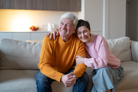 Affectionate senior wife hugs laughing husband as couple sits together on couch at home, joyful elderly spouses share humor, enjoy conversation and maintain close bond during retirement.