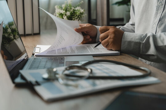 Hands reviewing and turning medical billing paperwork beside laptop and stethoscope, showing healthcare billing review, insurance claim checking and document workflow.