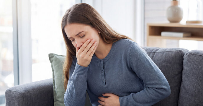 Sick young woman experiencing sudden nausea and severe stomach pain while sitting indoors on a comfortable living room couch