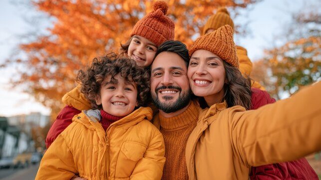 A joyful family of four wearing warm autumn clothes takes a selfie outdoors among colorful fall foliage.