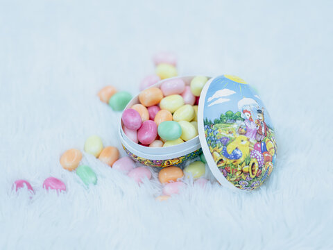 Pastel colored Easter candies spilling from an open decorative egg-shaped container on a soft white fluffy background.